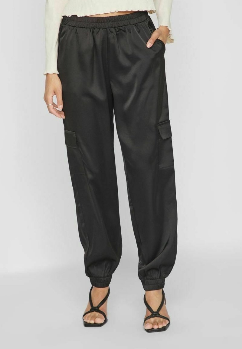 Femme portant un pantalon cargo en satin noir avec des poignets élastiques, associé à des talons noirs à lanières et un haut court crème à manches longues.
