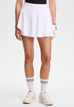 Donna che indossa una gonna da tennis bianca a strati, calzini bianchi con strisce nere, scarpe da ginnastica bianche e un guanto da golf bianco a una mano.