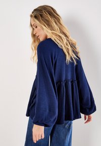 Navyblaues Oberteil mit Waffelstruktur, langen Glockenärmeln und gerafftem Taillenbereich, kombiniert mit lässigen Jeans.