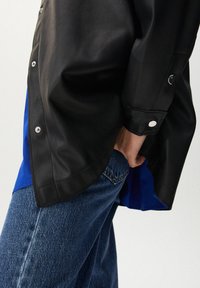 Person i blå jeans och en svart läderjacka med silverfärgade tryckknappar, handen instoppad i bakfickan på jeansen.