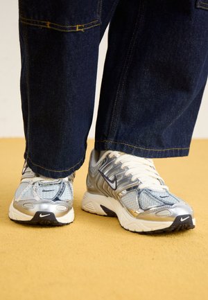 Baskets Nike argentées et blanches avec logos swoosh noirs portées avec un jean en denim bleu foncé sur un sol jaune.