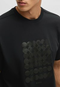 Sort kortærmet T-shirt med et tekstureret grafisk design bestående af cirkler i forskellige størrelser, nogle med striber, og med et brandingelement nederst.