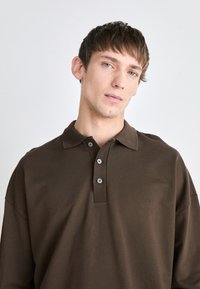Brun polo skjorte laget av mykt stoff, med en tre-knappers placket, korte ermer og en subtilt brodert logo på brystet.