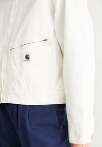 Vit bomullsjacka med en zip-fick på bröstet, logotyp-patch och en avslappnad passform. Kombineras med marinblå byxor.
