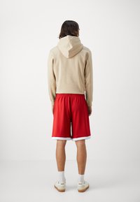 Sweat à capuche beige, short de sport rouge avec bordure blanche, et chaussures légères ; le tissu est lisse avec une taille élastique détaillée.