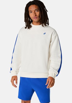 Mann mit Dreadlocks trägt weißen Pullover mit blauen Streifen und blaue Shorts, steht vor schlichtem, hellem Hintergrund.