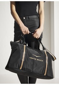 Black nylon duffel bag with gold accents and the text "VENICE BEACH." Features dual handles and a spacious main compartment.