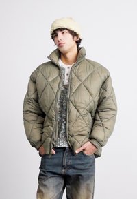 Olive green quilted puffer jacket with a high collar and front zipper, layered over a textured gray sweater, paired with faded jeans.