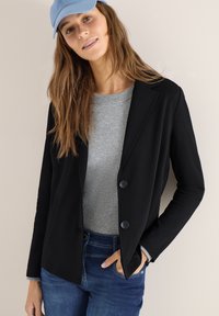 Femme portant un blazer noir sur un pull gris, un jean bleu et une casquette bleu clair, debout avec une main dans sa poche.