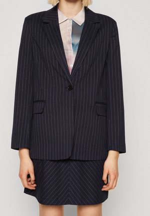 Frau trägt ein marineblaues Nadelstreifen-Blazer und einen passenden Rock über einem pastellfarbenen Hemd, stehend vor einem schlichten hellen Hintergrund.