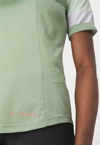 Vaaleanvihreä urheilupaita, jossa on teksturoitu kangas, lyhyet hihat ja valkoinen yksityiskohta. Helman lähellä on vaaleanpunainen "Vaude"-logo.