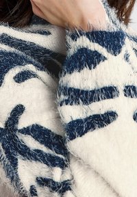 Écharpe en tricot texturé couleur crème avec motifs abstraits bleu foncé. Présente un matériau doux et duveteux avec des bords à franges pour plus de détails.