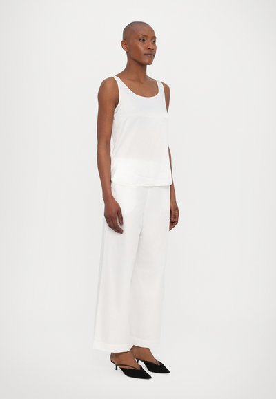 Valkoinen hihaton paita, jossa on pyöreä pääntie, yhdistettynä leveälahkeisiin valkoisiin housuihin ja mustiin korkokenkiin, luoden minimalistisen asun.