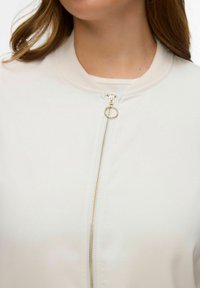 Blouson aviateur léger crème à texture lisse, doté d'une fermeture éclair dorée avec tirant en anneau, col côtelé et sans motifs visibles.