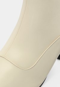 Gros plan d'une chaussure à talon haut en cuir beige lisse avec des détails de couture courbes et un bord de semelle noire visible au talon.