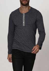 Tumman sininen pitkähihaine paita pehmeästä kankaasta, jossa on pyöreä kaula-aukko ja erottuva napitus etuosassa, sekä raidallinen kuvio keskiosassa.