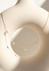 Collier en chaîne dorée avec des charms en forme de papillon ornés de pierres précieuses transparentes, exposé sur un mannequin minimaliste beige contre un fond clair.