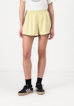 Model draagt gele sportshorts met bruine bies, zwarte sokken en zwarte Adidas sneakers met witte strepen en gumzolen.