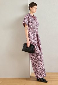 Rózsaszín leopárdmintás ruha rövid ujjal, deréknál övvel és oldalsó hasítékkal. Fekete táskával és bokacsizmával párosítva. Fa padló háttér.