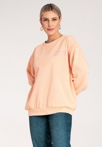 Brzoskwiniowy, oversize'owy sweatshirt z okrągłym dekoltem i ściągaczem na dole. Posiada haftowany napis na piersi w jasnofioletowym kolorze. Miękka tekstura materiału.