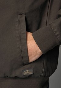 Jachetă maro închis dintr-un material texturat. Are un buzunar lateral și un mansetă cu arici. Eticheta de lângă buzunar afișează detalii despre marcă și logo-ul.