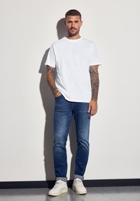 Bílá bavlněná trička, modré džíny s rovný fit a bílé tenisky. Model má na rukou tetování. Minimalistický design, ležérní styl.