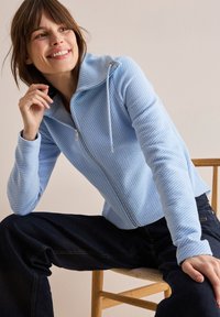 Giacca zip-up azzurro chiaro realizzata in tessuto testurizzato con cappuccio, caratterizzata da un leggero motivo diagonale e maniche lunghe, abbinata a jeans scuri.