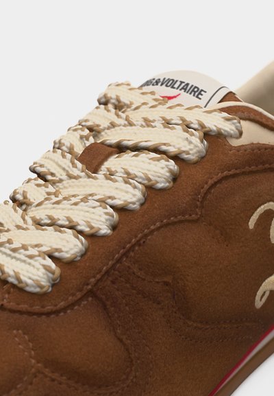 Καφέ σουέντ αθλητικό παπούτσι με χοντρά κρεμ και καφέ στριμμένα κορδόνια, ορατή ραφή και λευκή ετικέτα γλώσσας με μαύρο και κόκκινο κείμενο.