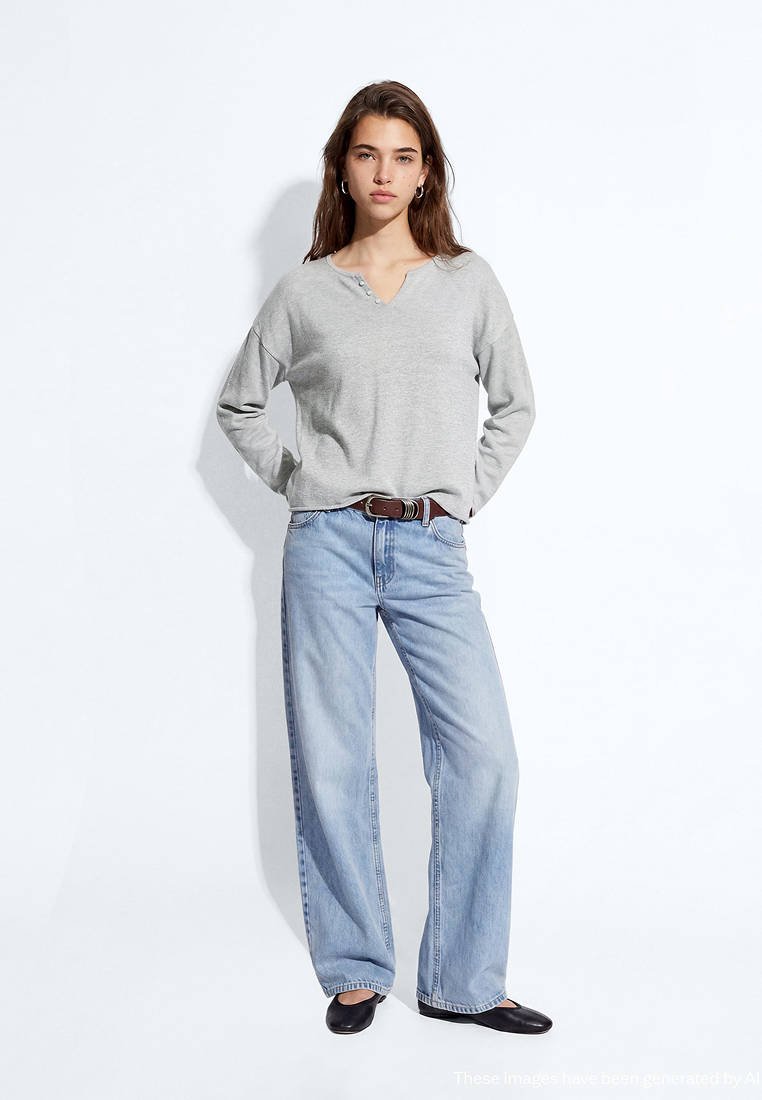 Jeune femme portant un haut à manches longues gris clair, un jean bleu clair ample avec une ceinture marron, et des chaussures plates noires, debout devant un fond blanc.