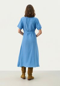 Lungo vestito in lino blu con maniche corte, cintato in vita, che scende fino a metà polpaccio, abbinato a stivali in suede marroni alti fino al ginocchio.
