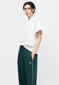 Νεαρός με κοντά σκούρα μαλλιά που φοράει λευκό φαρδύ μπλουζάκι και σκούρο πράσινο παντελόνι με φαρδιά πόδια και λευκή ρίγα, στέκεται με το ένα χέρι στην τσέπη.