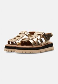 Gouden geweven sandaals met open teen, voorzien van een chunky zool met zwart en beige accenten. De bandjes zijn metallic, wat zorgt voor ondersteuning en stijl.