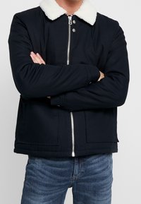 Svart jacka med vit pälskrage, dragkedjeslutning och två framfickor. Tyget verkar slätt med en strukturerad design.