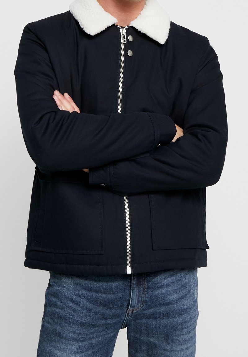 Svart jacka med vit pälskrage, dragkedjeslutning och två framfickor. Tyget verkar slätt med en strukturerad design.