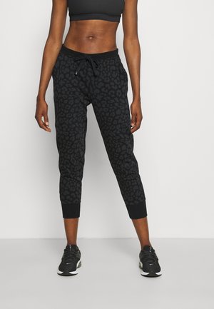 Kvinde iført sorte korte leopardmønstrede joggingbukser, sort sports-bh og sorte løbesko, stående mod en ensartet lys baggrund.