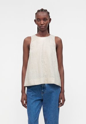 Γυναίκα με πλεξούδες φοράει αμάνικη μπεζ μπλούζα και μπλε τζιν, στέκεται μπροστά σε ένα απλό φωτεινό φόντο.