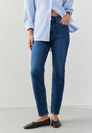 Frau trägt blaue Slim-Fit-Jeans, schwarze Ballerinas und ein hellblau gestreiftes, geknöpftes Hemd, eine Hand in der Tasche.