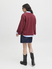 Bordeauxroter übergroßer Sweatshirt mit langen Ärmeln, über einem hellblauen Hemd mit Kragen getragen, kombiniert mit einem dunklen Jeansrock und schwarzen Stiefeln bis zum Knie.