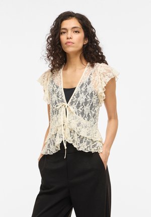 Vrouw met krullend haar, gekleed in een crèmekleurige kanten blouse met korte mouwen, die aan de voorkant is gestrikt over een zwart topje, gecombineerd met een zwarte broek, handen in de zakken.