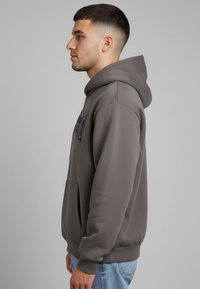 Noor mees halli kapuutsiga pusa ja siniste teksadega, vaadates vasakule, ühevärvilise halli tausta ees.