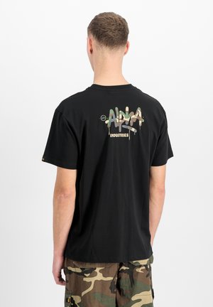 Man wearing black T-shirt with camo-style "Alpha Industries" logo on back and camo cargo shorts, standing against white background.