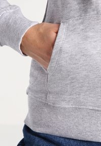 Sudadera gris con dobladillo y puños de canalé, que presenta un bolsillo lateral. La tela parece suave y texturizada, apta para el uso diario.