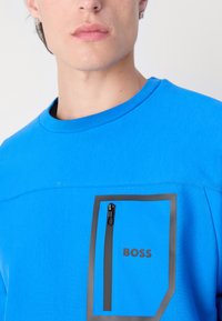Felpa blu realizzata in un materiale leggero e testurizzato. Presenta una tasca con zip a contrasto con bordo nero e il logo del brand stampato.