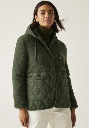 Mujer vistiendo una chaqueta híbrida acolchada y de forro polar en verde con capucha y bolsillos frontales, combinada con pantalones beige sobre un fondo neutro.