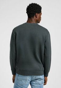 Felpa grigia scura con colletto a girocollo, maniche lunghe, polsini e orlo a costine, e una texture liscia. Indossata con jeans azzurri chiari.