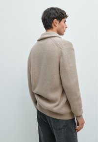 Beige Strickpullover mit Kragen und gerippter Textur, mit lässigen Schultern und lockerer Passform, kombiniert mit dunkelgrauen Jeans.