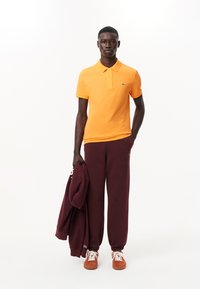 Orange polo shirt med korta ärmar, i kombination med bourgognefärgade sweatpants. Modellen håller en matchande bourgogne hoodie. Orange sneakers fulländar outfiten.