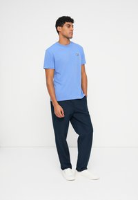 Világoskék, rövid ujjú póló, puhára kidolgozott anyagból, kis logóval. Tengerészkék nadrággal és fehér sportcipővel párosítva, hogy kontrasztot képezzen.