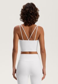 White athletic crop top with thin, crisscross back straps and smooth, stretchy fabric. Worn with matching white high-waisted leggings.