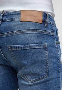 Jeans de mezclilla azul oscuro con un parche de cuero etiquetado como "ZY PREMIUM". Las características incluyen cinco bolsillos y detalles de costura marrón.